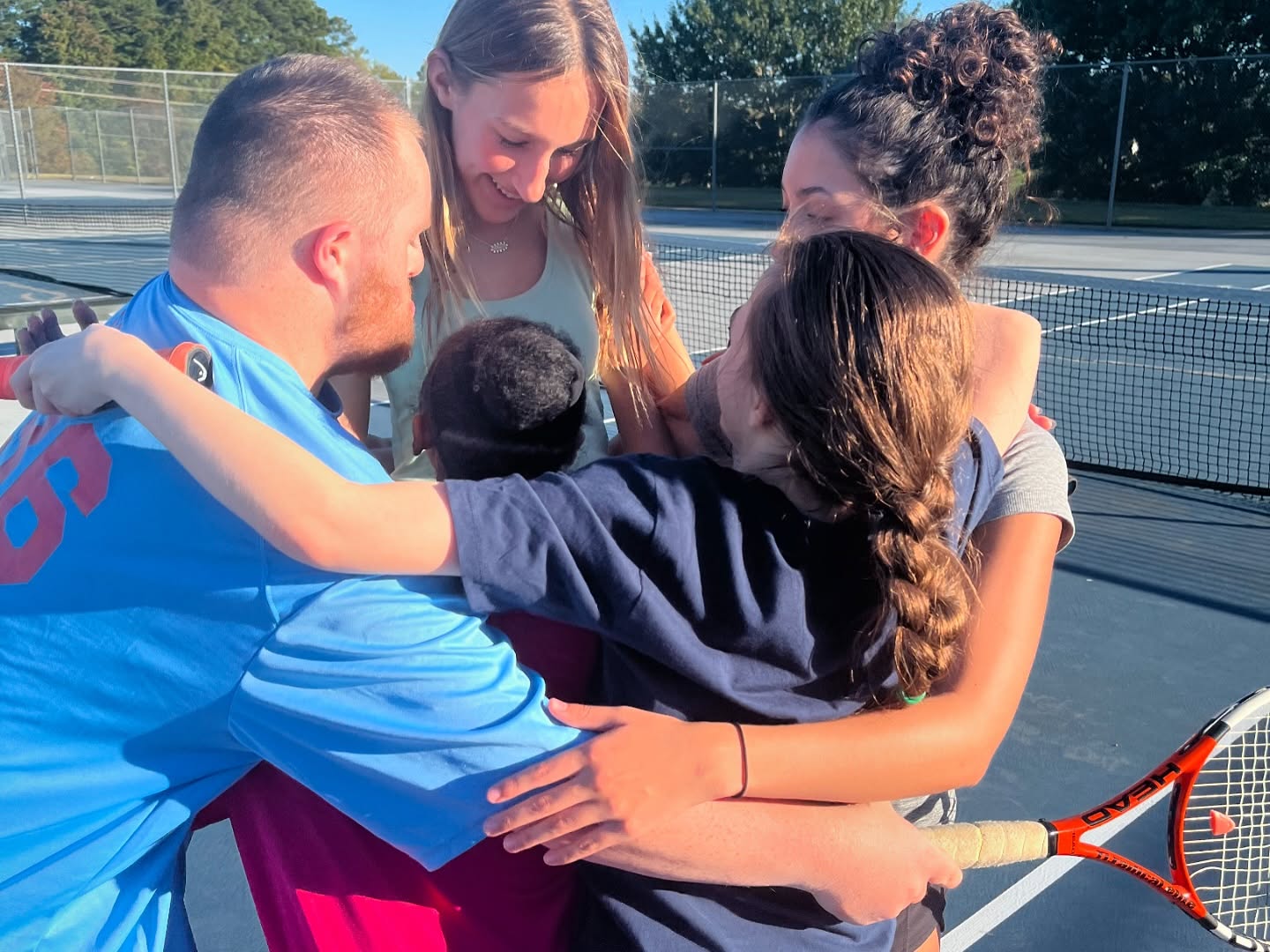 A group of five people hugging on a tennis court, with a sunny background and a tennis racket visible.