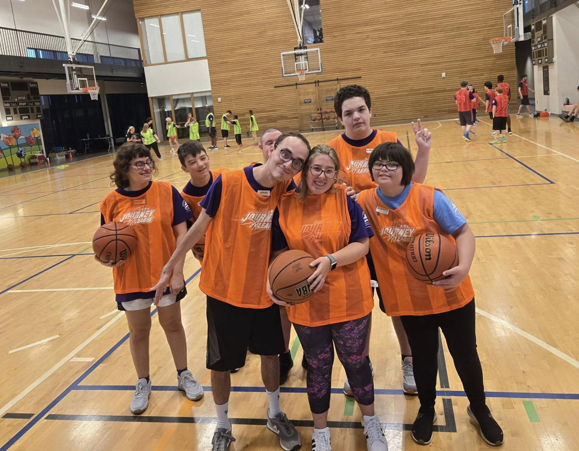 A group of six people in orange vests smiling and holding basketballs in a gym.
