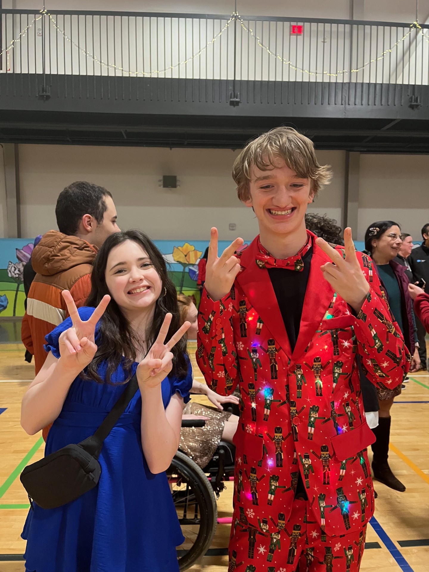 Two people smiling and showing peace signs, one in a blue dress and the other in a red nutcracker-patterned suit, in a gymnasium setting.