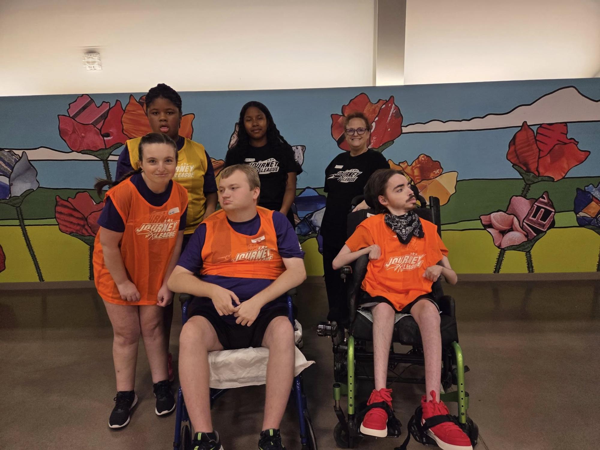 A group of six people posing in front of a colorful floral mural, with some wearing "Journey League" jerseys.